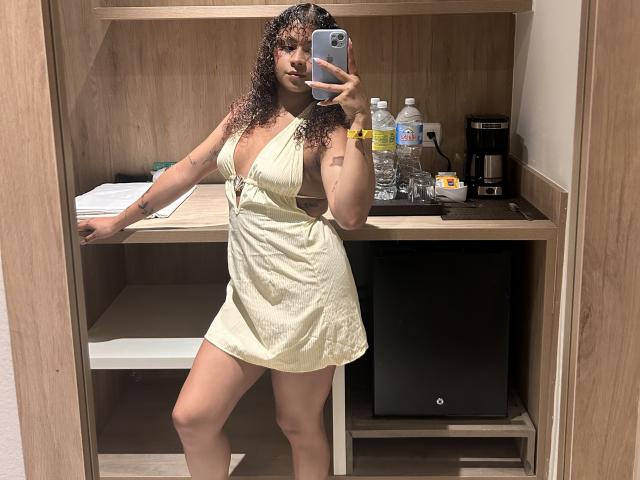 SofiaPassionne with curly hair, wearing a yellow dress with side cutouts, showing her tits, holding a phone, standing in a mirror, in a room with a wooden desk.