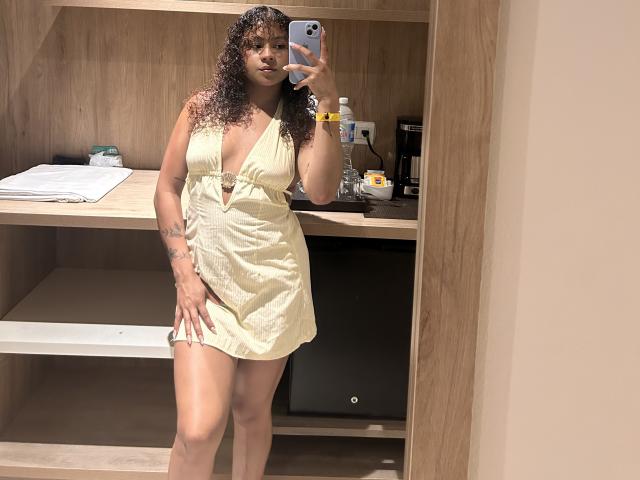 SofiaPassionne with curly brown hair, tan skin, small tattoos, wearing a yellow, deep-strap dress, holding a phone, taking a mirror selfie, showing her ass.
