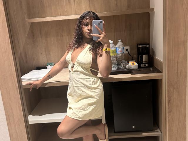 SofiaPassionne with curly brown hair, yellow dress with cutouts, holding phone, showing her tits, standing in front of wooden mirror, water bottle, coffee maker, and shelf.