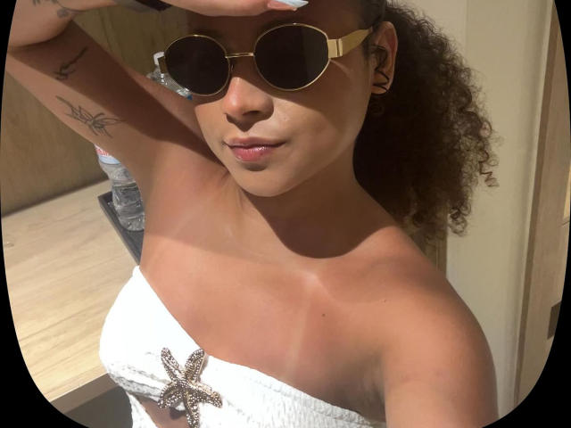 SofiaPassionne with curly brown hair, tan skin, wearing gold aviator sunglasses, white strapless top with a star-shaped jewel, and a subtle smile, showing her small, perky tits.