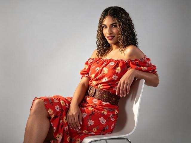 StefannyMia with curly brown hair, red off-shoulder floral dress, brown belt, sitting on white chair, smiling, hands on chair, grey background, visible ass, cute, sexy.