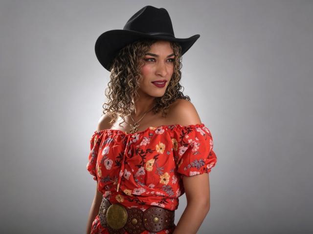 StefannyMia with curly brown hair, wearing a black cowboy hat, red floral off-shoulder top, and brown belt, smiling at the camera, showcasing her tits and cute face.