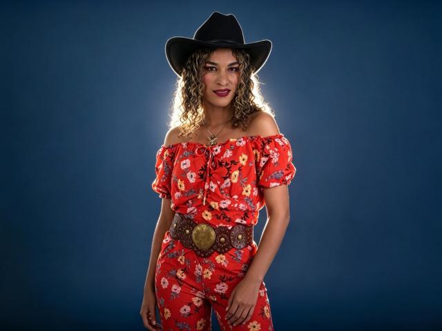StefannyMia in a red floral off-shoulder top and pants, black cowboy hat, curly hair, dark lipstick, big brown belt with gold buckle, blue background, smiling.