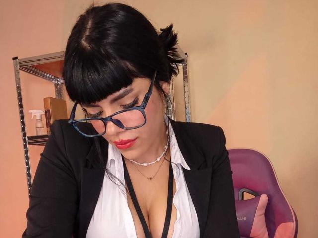 AfroditaMooreX, con capelli neri, occhiali e rossetto rosso, indossa un blazer nero e una camicia bianca, mostrando il décolleté, in una stanza con una sedia viola e una mensola..