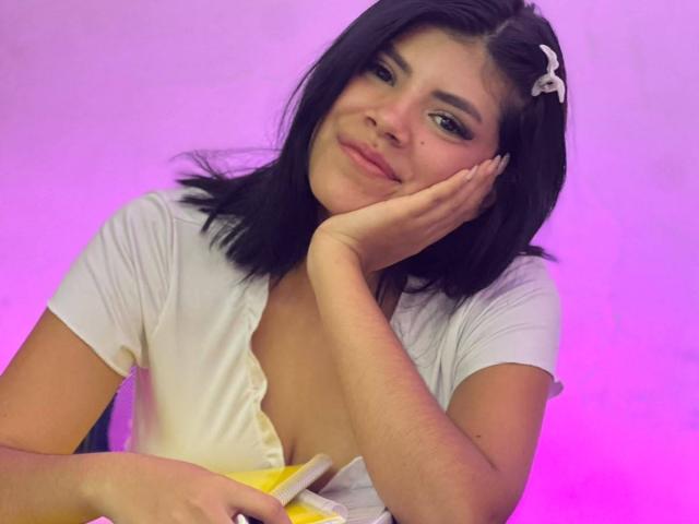 AfroditaMooreX with black hair, white shirt, and pink background, resting her cheek on her hand, holding a yellow object, smiling subtly, small white hairclip in her hair.