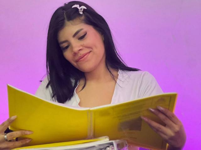 AfroditaMooreX, avec des cheveux noirs, une chemise blanche et une pince à cheveux rose, lisant un livre jaune, souriant doucement, tenant le livre ouvert avec les deux mains, fond rose-violet..