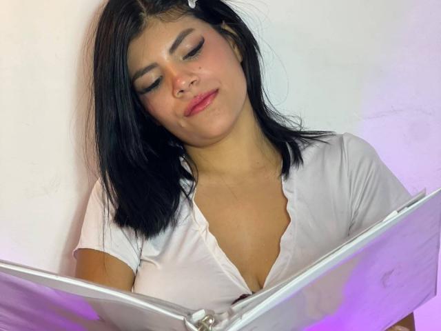 AfroditaMooreX with black hair, white shirt, and pink lipstick, eyes closed, slightly smiling, holding open white book with purple light, showing her small tits and cleavage.