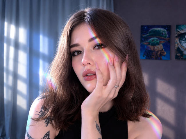 TrudieProse with brown hair, tattoos on arms, hand on face, rainbow light on cheek, wearing black top, soft expression, light from window, blue curtains in background.