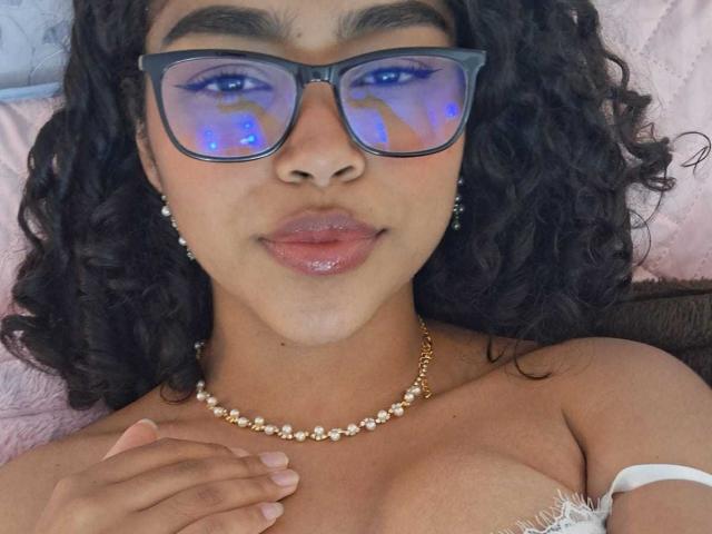 FernandaCoa with dark curly hair, wearing glasses with blue light reflection, pearl necklace, and white strap, touching her chest, glossy pink lips, and light brown skin.