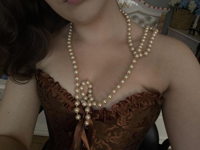 Dreia's tits in a brown lace corset, showing off a long pearl necklace that rests between her cleavage. Her brown hair is partially visible.