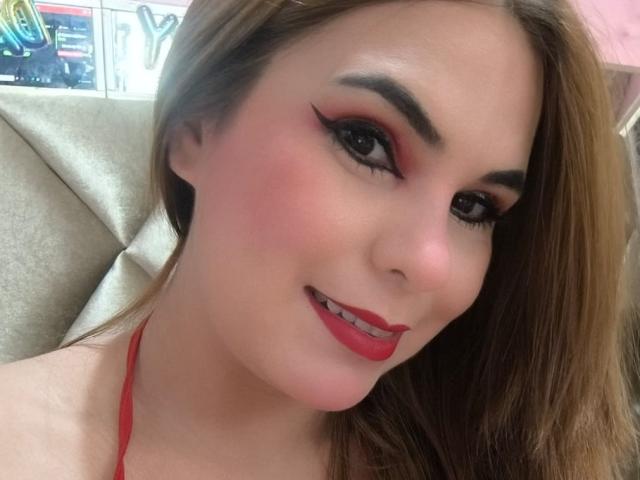 AuroraRossi has red lipstick, pink blush, and thick black eyeliner. She's smiling with red-clipped hair and a red halter top. Her tits are slightly visible.