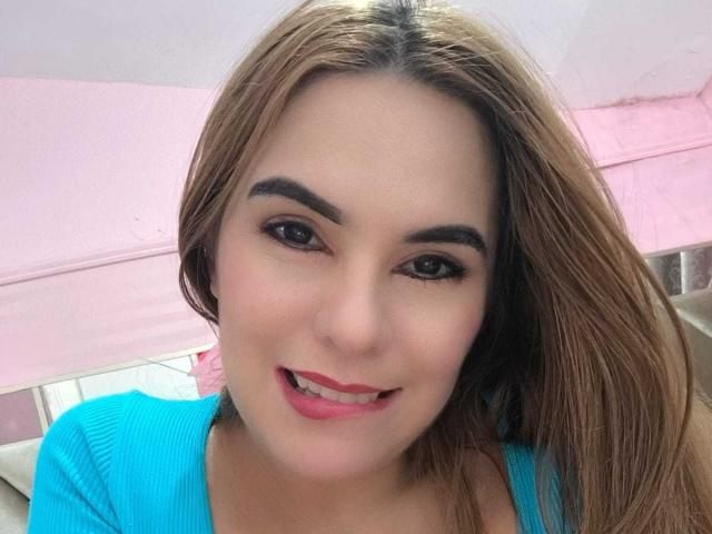 AuroraRossi has light brown hair, dark eyebrows, brown eyes, pink lipstick, and a cute smile wearing a blue top, with a slightly parted red mouth.
