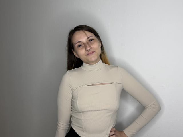 SharaPiette, com cabelo castanho claro, gola alta bege, seios pequenos, mãos na cintura, sorrindo, em pé contra uma parede branca, enquadramento frontal, pele clara, casual, confiante..