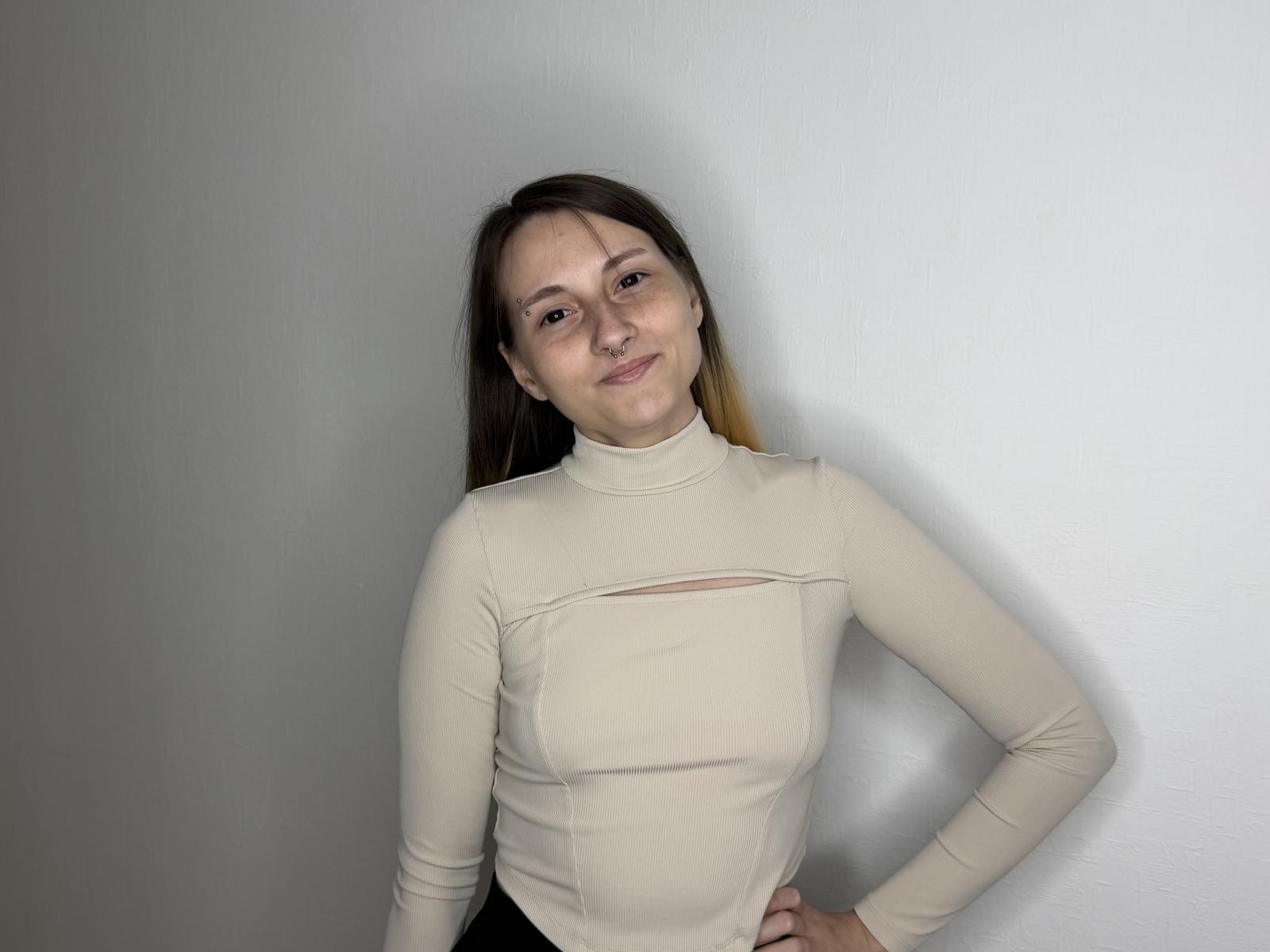 SharaPiette mit hellbraunen Haaren, einem beigen Rollkragenpullover, kleinen Brüsten, mit den Händen an den Hüften, lächelnd, stehend vor einer weißen Wand, frontal, helle Haut, lässig, selbstbewusst..