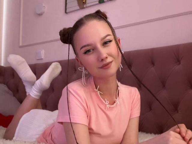 KizzieWickson with double buns, pink shirt, white socks, necklace, holding hair, lying on bed, cute, smiling, pink room, tufted headboard, white pillow, relaxed, soft lighting.