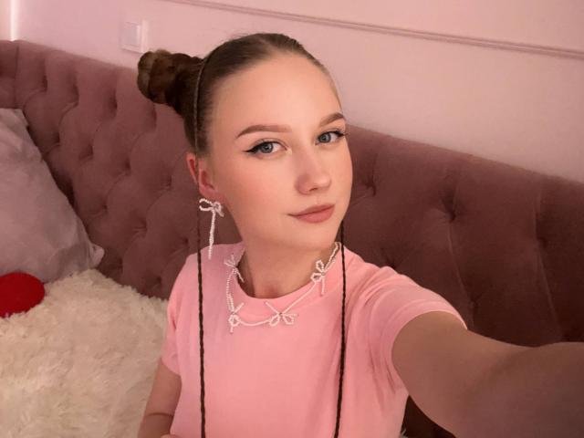 KizzieWickson with braided pigtails, wearing a pink shirt, sitting on a pink tufted couch, smiling at the camera, her tits are small, blue eyes, light skin.