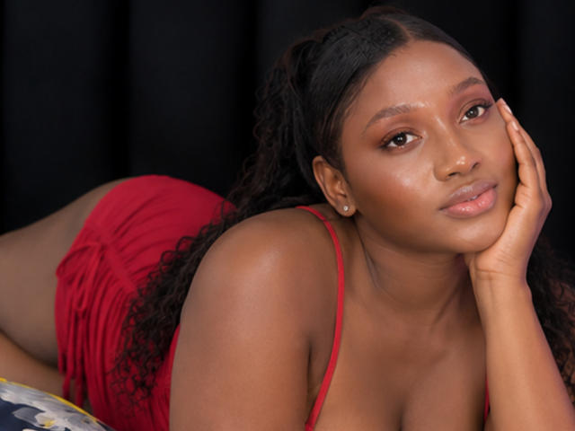CocoaBell has dark skin, curly black hair, and wears a red dress. She's lying on her side, resting her head on her hand, with a soft, sexy smile.