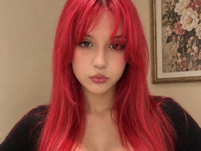 ScarletMyst with bright red hair, nose piercing, and glossy pink lips, wearing a black top, staring directly at the camera with a neutral expression.