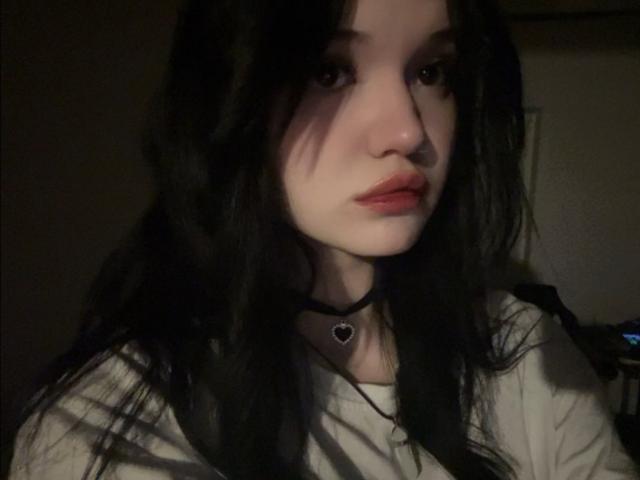 MistyEllstrom ha la pelle chiara, i capelli neri e ondulati, le labbra rosse e indossa una collana nera con un ciondolo a forma di cuore, in una foto con una luce fioca. La sua espressione è leggermente corrucciata..