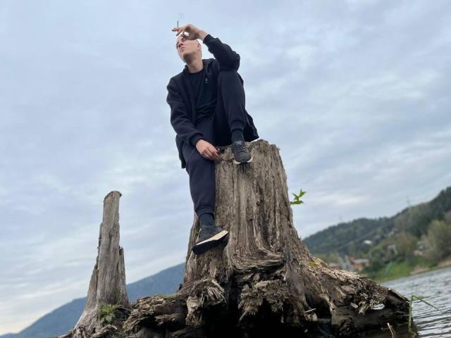 MisterHardcore sits on a decaying tree stump, pointing up with his right hand, wearing a black hoodie and sneakers, with overcast sky and water in background.