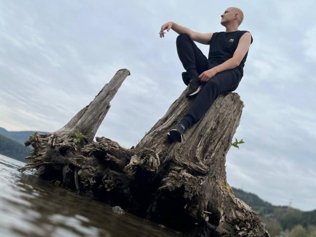 MisterHardcore est assis sur un tronc d'arbre mort et rugueux, dans l'eau, vêtu de vêtements noirs et de baskets, avec une tête chauve et une posture détendue et confiante..