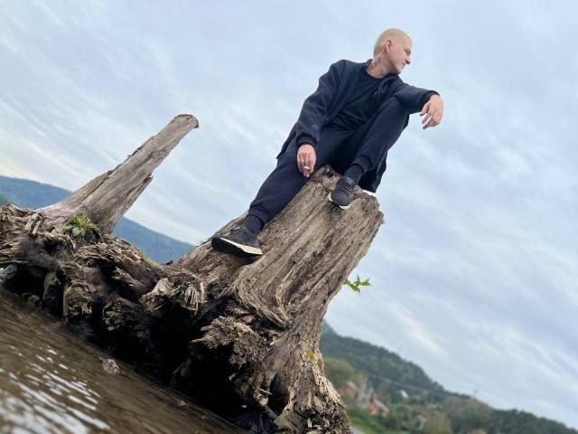 MisterHardcore, calvo, vestito di nero, seduto su un tronco di legno grezzo vicino all'acqua, con la gamba destra sollevata, il braccio sinistro appoggiato, che guarda di lato, cielo nuvoloso, giornata nuvolosa..