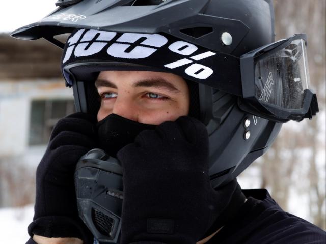 MisterHardcore mit blauen Augen, der einen schwarzen Helm, schwarze Handschuhe und eine schwarze Maske trägt, berührt sein Gesicht und zeigt seine Nase und Augen, in einer verschneiten Outdoor-Umgebung..
