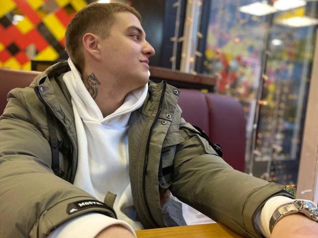 MisterHardcore mit kurzen, braunen Haaren, einer grünen Jacke, einer weißen Kapuzenjacke, einem Tattoo am Hals, der zur Seite schaut, einer Armbanduhr, einem Holztisch, einer roten Sitzgruppe, einem ungezwungenen Restaurant, mit konzentriertem Gesichtsausdruck..