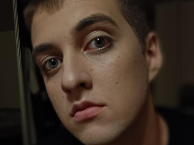 Le visage de MisterHardcore, cheveux bruns courts, yeux verts, peau claire, légère imperfection sur la joue droite, expression neutre, lèvres rosées légèrement entrouvertes, arrière-plan sombre flou..