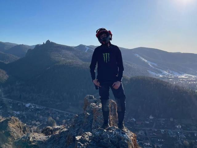 MisterHardcore se tient sur un sommet rocheux, portant un t-shirt noir Monster, des lunettes de protection et un casque rouge, avec des montagnes enneigées et une ville en contrebas, en plein jour, les mains le long du corps, regardant vers l'avant..