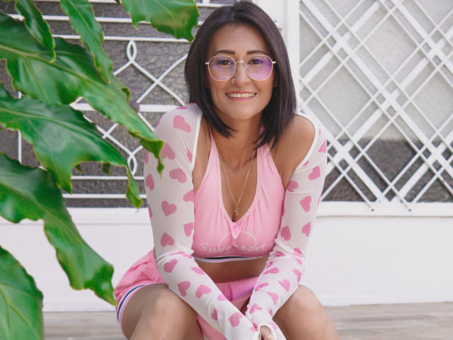 SharissWilliam with pink heart-patterned sleeves, pink crop top, and pink shorts, sitting, smiling, wearing pink glasses, and green leaves in the foreground. She has a cute, slim, and sexy body.