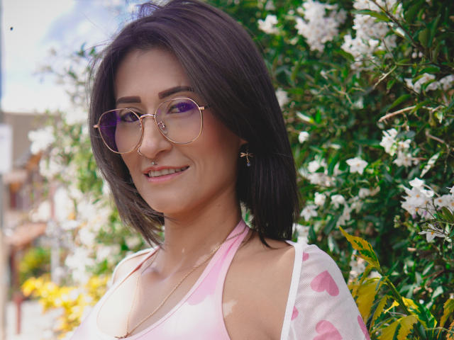 SharissWilliam with dark brown hair, pink glasses, and a pink polka dot top, smiling, showing her tits slightly, surrounded by white flowers and green leaves.