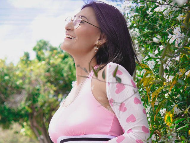 SharissWilliam with big tits, wearing pink heart-patterned sleeves, and pink top, smiling in sunlight, has dark hair, glasses, and gold earrings, standing by green, leafy bushes.