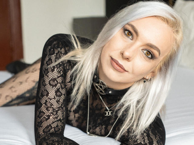VelvetTanne with blonde hair, black lace top, and a choker, lying on a bed, looking at the camera with brown eyes, wearing a necklace, smiling slightly.
