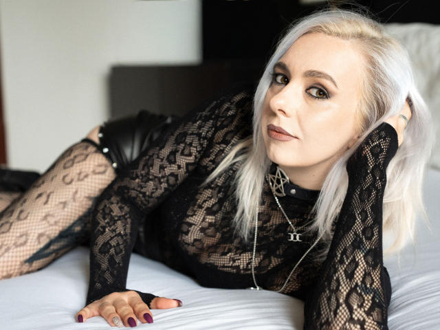 VelvetTanne with white hair, black lace outfit, and leather pants, lying on bed, looking at camera, hand in hair, purple nails, black choker with pentagram, confident smile.