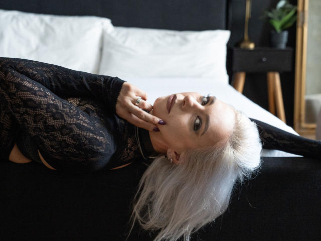 VelvetTanne with platinum blonde hair, black lace top, lying on bed, hand near face, seductive gaze, black headband, white pillows, black bed, ring on finger, dark nails.