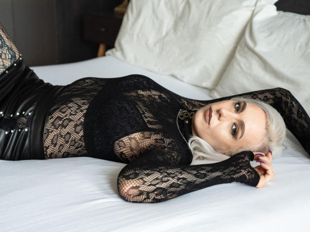 VelvetTanne lying on a bed, wearing a black lace top, black leather pants, and a choker, with pale skin and white hair, looking at the camera.
