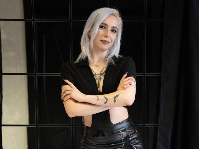 VelvetTanne with white hair, black shirt, tattoos on arms and chest, leather pants, arms crossed, smiling, standing in front of black grid background.