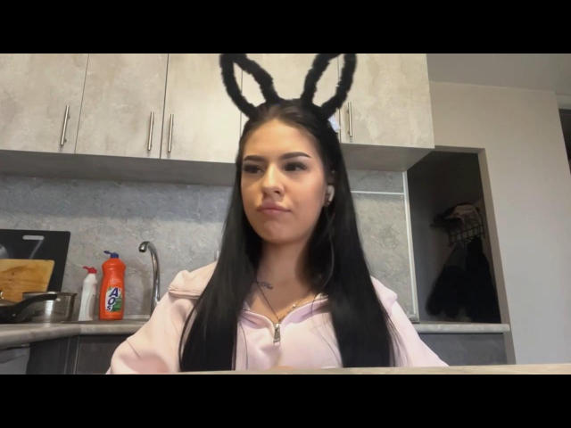 CorrieBogacz with long black hair, wearing bunny ears, pink hoodie, and a serious face in a kitchen, has a small tattoo on her chest and a nose ring.