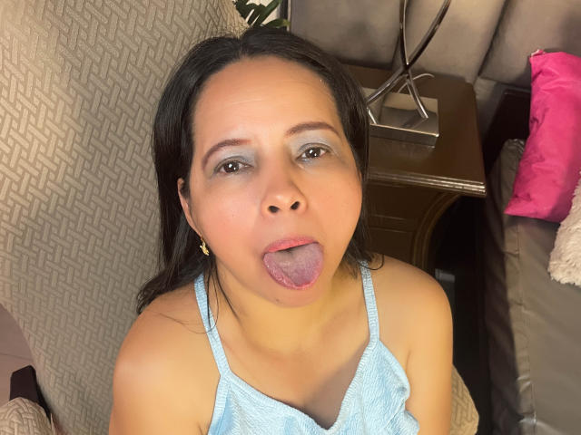 SelenaBeltran with tongue out, blue tank top, medium tits, brown eyes, light makeup, long black hair, beige patterned chair, pink pillow, wooden table, and metal lamp in background.