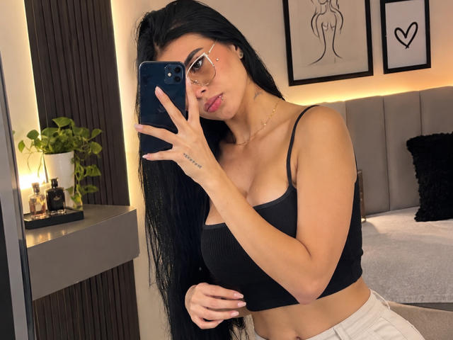 AmilyBella with long black hair, glasses, and a black crop top, holding a phone to her face, showing her tits and a small tattoo on her left hand.