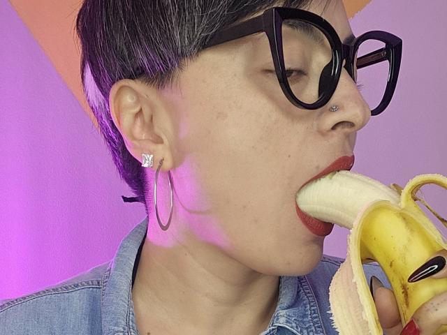 KatParadise with short black hair, black glasses, nose ring, and hoop earrings, eating a banana with red lipstick, wearing a denim shirt. KatParadise's face is slightly turned right.