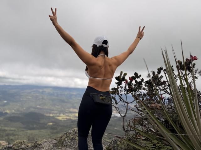 EmaRusso with arms raised, wearing a white cap, backless top showing her ass, and black pants, standing on rocky terrain with a "peace" sign, surrounded by greenery.