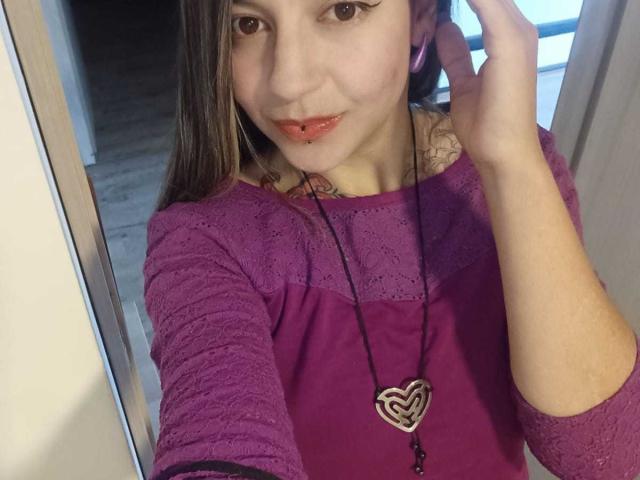 ArianaBlonde with fair skin, pink lips, and brown eyes, wearing a purple lace top, heart necklace, and pink earphones, smiling at the camera. Tattoo visible on her neck.