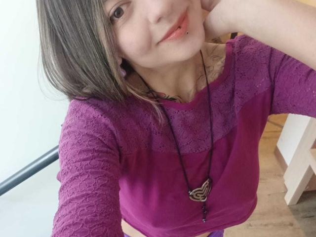 ArianaBlonde with a cute smile, pink lip ring, and brown hair, wearing a purple lace top, and a heart necklace, holding her head. Boobs are partially visible.