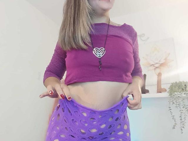 ArianaBlonde with pink top, purple mesh pants, showing her midriff, pulling pants down, heart necklace, blonde hair, red nails, in a bright room, slightly visible ass.