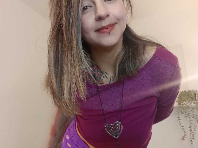 ArianaBlonde with light brown hair, purple top, heart necklace, and tattoo on chest, smiling with slightly bent over, showing small, perky tits and subtle ass.