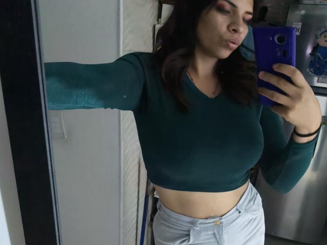 VioletaStone with big tits, wearing a green long-sleeve crop top and light gray pants, taking a mirror selfie with a blue phone, pouting her lips.
