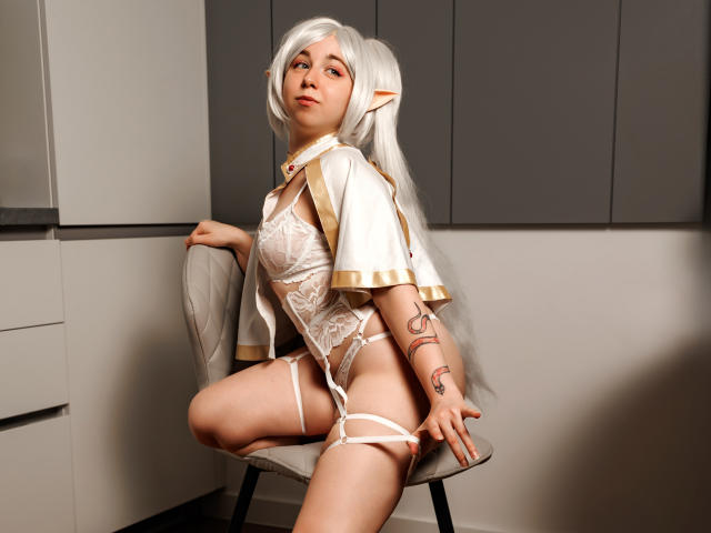 AlexxisSky with white cosplay outfit, lace lingerie, and garter, sitting on chair, exposing her pussy, white hair, elf ears, tattoos, and looking up. Bedroom-style setting, sexy.
