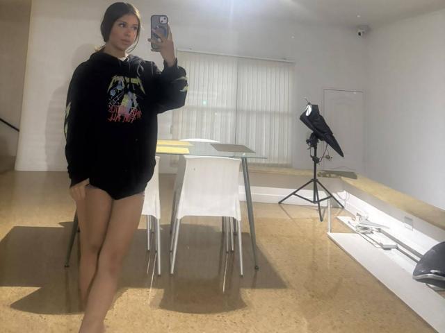 SalyFoster with long dark hair, wearing a black graphic sweatshirt and no pants, showing her ass, standing in a room with a mirror, holding a phone.